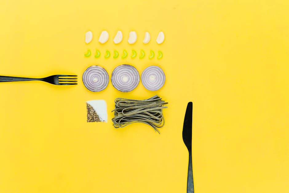 A colorful flatlay of pasta, onion, garlic, herbs, and cutlery on a yellow background.